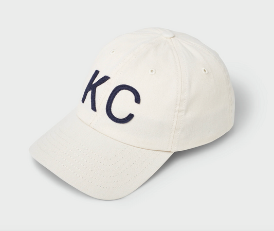 An off-white cotton baseball cap with dark navy blue embroidered letters 'KC' on the front, shown on a light gray background.
