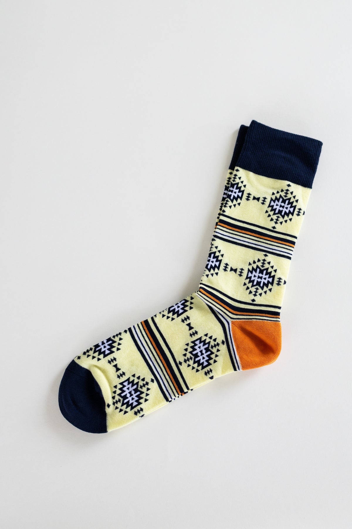A pair of patterned crew socks laid flat on a light gray background. The sock has a pale yellow base with navy blue geometric diamond motifs, navy blue cuffs and toes, and horizontal stripes in navy, orange, and thin beige lines.