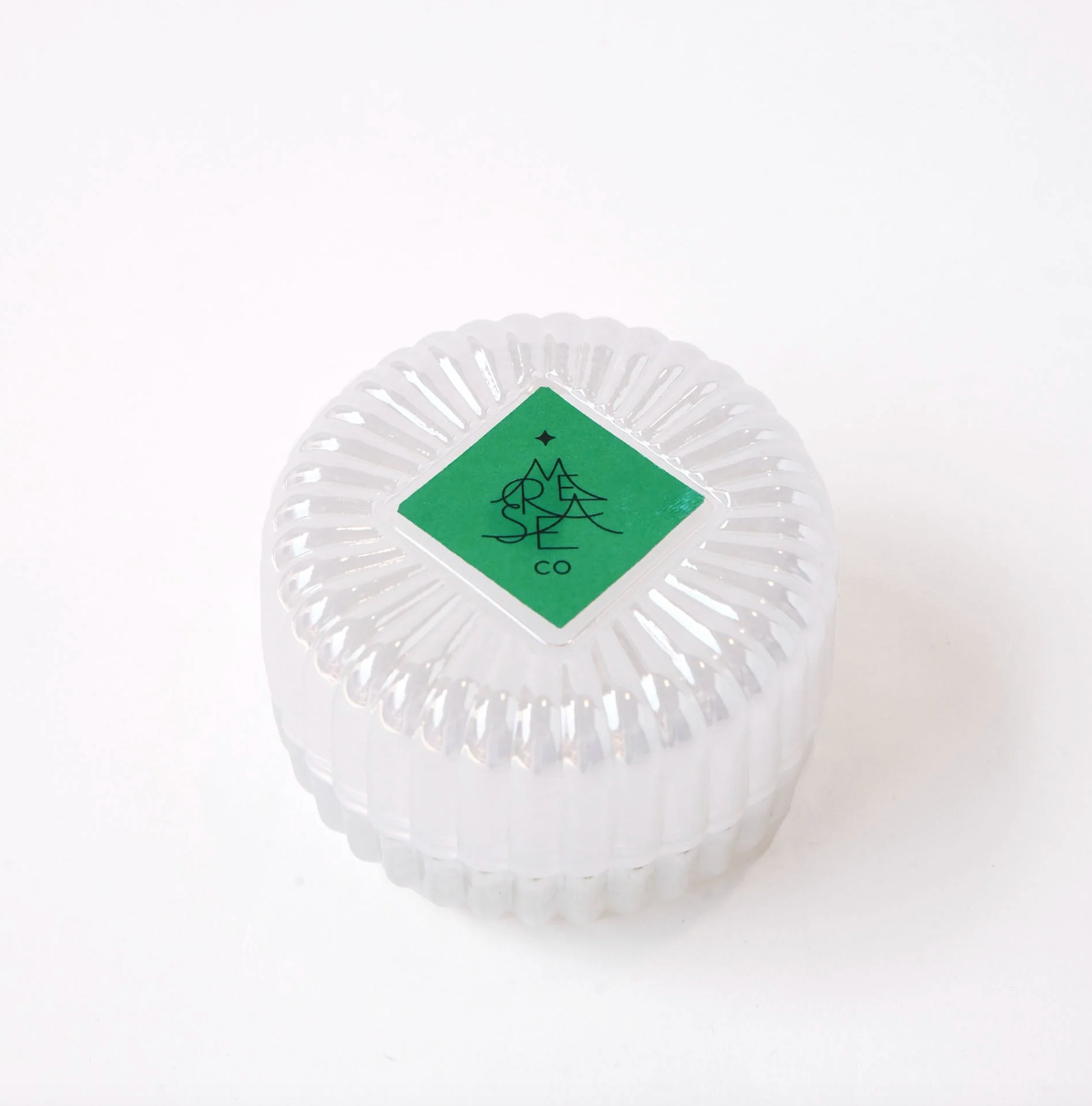 A clear ribbed glass round container with a green diamond-shaped label on top featuring a black symbol and the text 'MERSEA Co'.