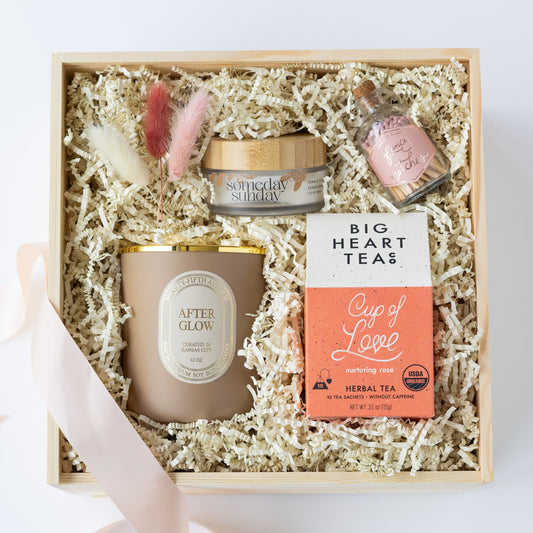 A curated gift box set on a white background, containing a beige candle, a small pink bottle of matches, a Big Heart Tea Cup of Love tea box, and a jar of Someday Sunday Body Lotion, all nestled in white paper krinkle inside a light wood box with a pale pink ribbon.