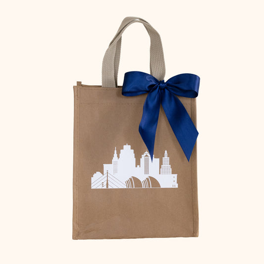 A brown paper gift bag with beige handles, decorated with a white Kansas City skyline graphic and a large navy blue satin bow, shown against a solid cream background.