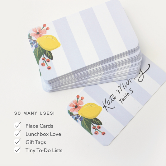 A stack of note cards with a white and light blue striped design, decorated with a yellow lemon, pink flowers, and green leaves. One card is shown flat with a printed example name and table number.