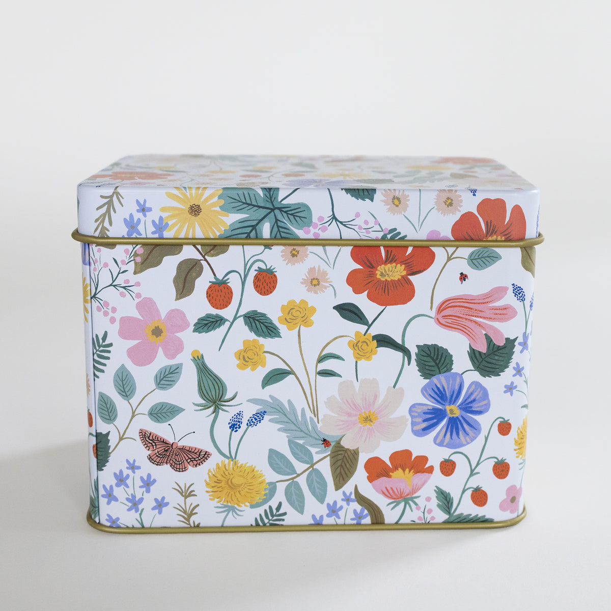 A square metal tin with a colorful floral and botanical print on a white background, featuring pink, red, yellow, blue, and green flowers with leaves, berries, and a butterfly. The tin has a gold-toned metal rim and is photographed against a clean, light off-white background.
