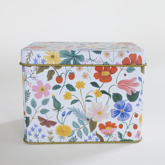 A square metal tin with a colorful floral and botanical print on a white background, featuring pink, red, yellow, blue, and green flowers with leaves, berries, and a butterfly. The tin has a gold-toned metal rim and is photographed against a clean, light off-white background.