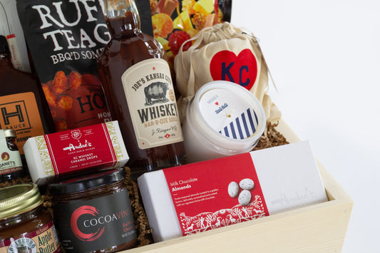 A wooden gift box filled with assorted gourmet Kansas City snacks, tea, coffee, and packaged food items, photographed against a clean white background.