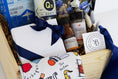 Load image into Gallery viewer, A curated wooden gift box filled with assorted Kansas City sourced packaged goods, including a white chocolate box tied with a navy ribbon, Jack Stack seasoning and bbq sauce, a blue Roasterie coffee bag, a printed Kansas City themed towel, a barbecue sauce bottle from Q39, and a Kansas City Royals blue edition of Topsy's popcorn mix, photographed in a clean studio setup.
