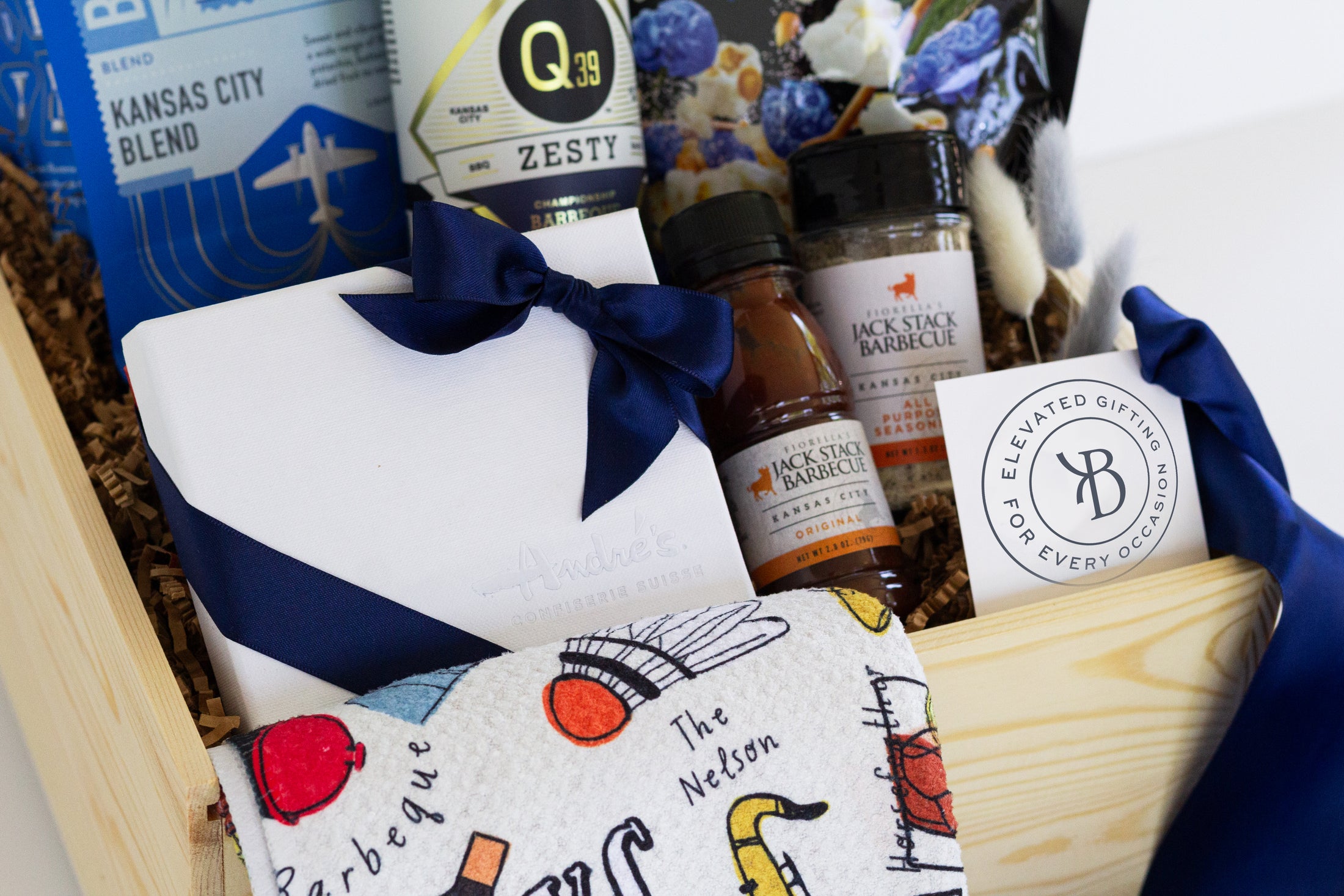A curated wooden gift box filled with assorted Kansas City sourced packaged goods, including a white chocolate box tied with a navy ribbon, Jack Stack seasoning and bbq sauce, a blue Roasterie coffee bag, a printed Kansas City themed towel, a barbecue sauce bottle from Q39, and a Kansas City Royals blue edition of Topsy's popcorn mix, photographed in a clean studio setup.