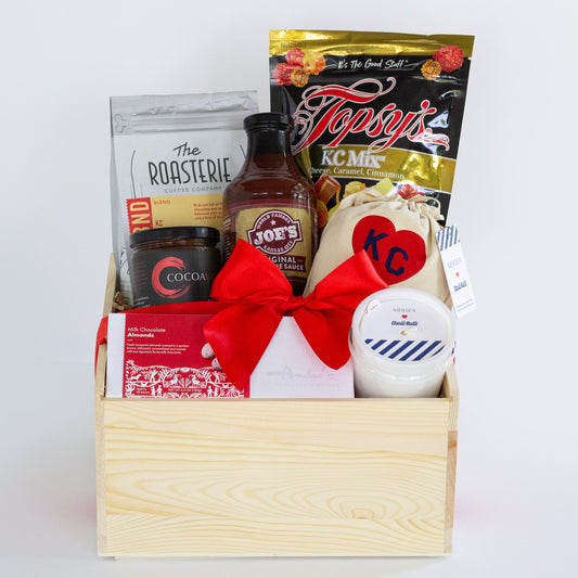 A wooden gift basket filled with assorted food items including a bag of Topsy's Popcorn KC Mix, a package of The Roasterie Coffee, a jar of Cocoavino, Joe's BBQ Sauce, a KC Heart Candle displayed in and outside of the candle pouch, and Andre's chocolates, tied with a red ribbon on a white background.