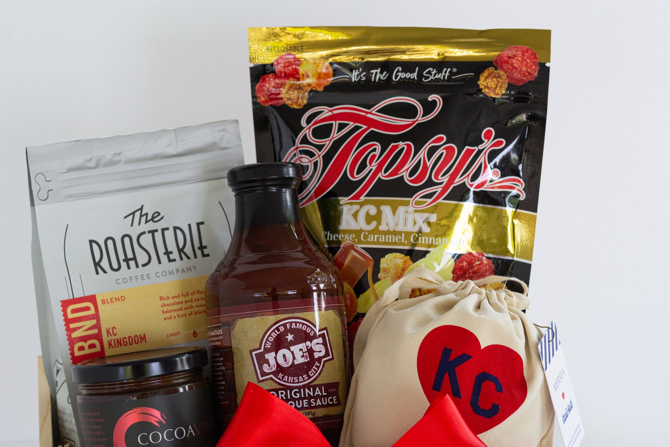 A wooden gift basket filled with assorted food items including a bag of Topsy's Popcorn KC Mix, a package of The Roasterie Coffee, a jar of Cocoavino, Joe's BBQ Sauce, a KC Heart Candle displayed in and outside of the candle pouch, and Andre's chocolates, tied with a red ribbon on a white background.