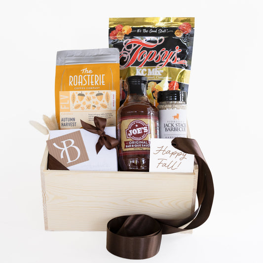 A fall-themed gourmet gift basket in a light wood crate, filled with various food items including a bag of The Roasterie Autumn Harvest Blend, a bottle of Joe's Originals BBQ Sauce, a bag of Topsy's Popcorn KC Mix and a handwritten 'Happy Fall!' card, accented with a dark brown ribbon.