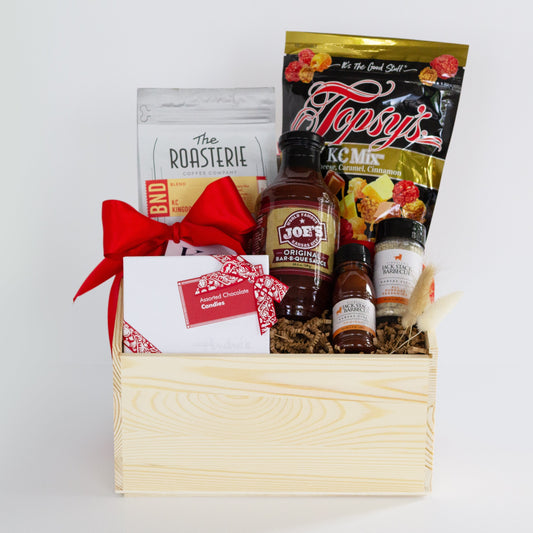 A wooden gift basket filled with coffee and tea items, including Andre's Chocolates, a bag of Topsy's Popcorn KC Mix, Roasterie Coffee, Joe's BBQ Sauce, and Jack Stack mini seasoning and bbq sauce set, arranged in a light wood crate with a red ribbon on a white background.