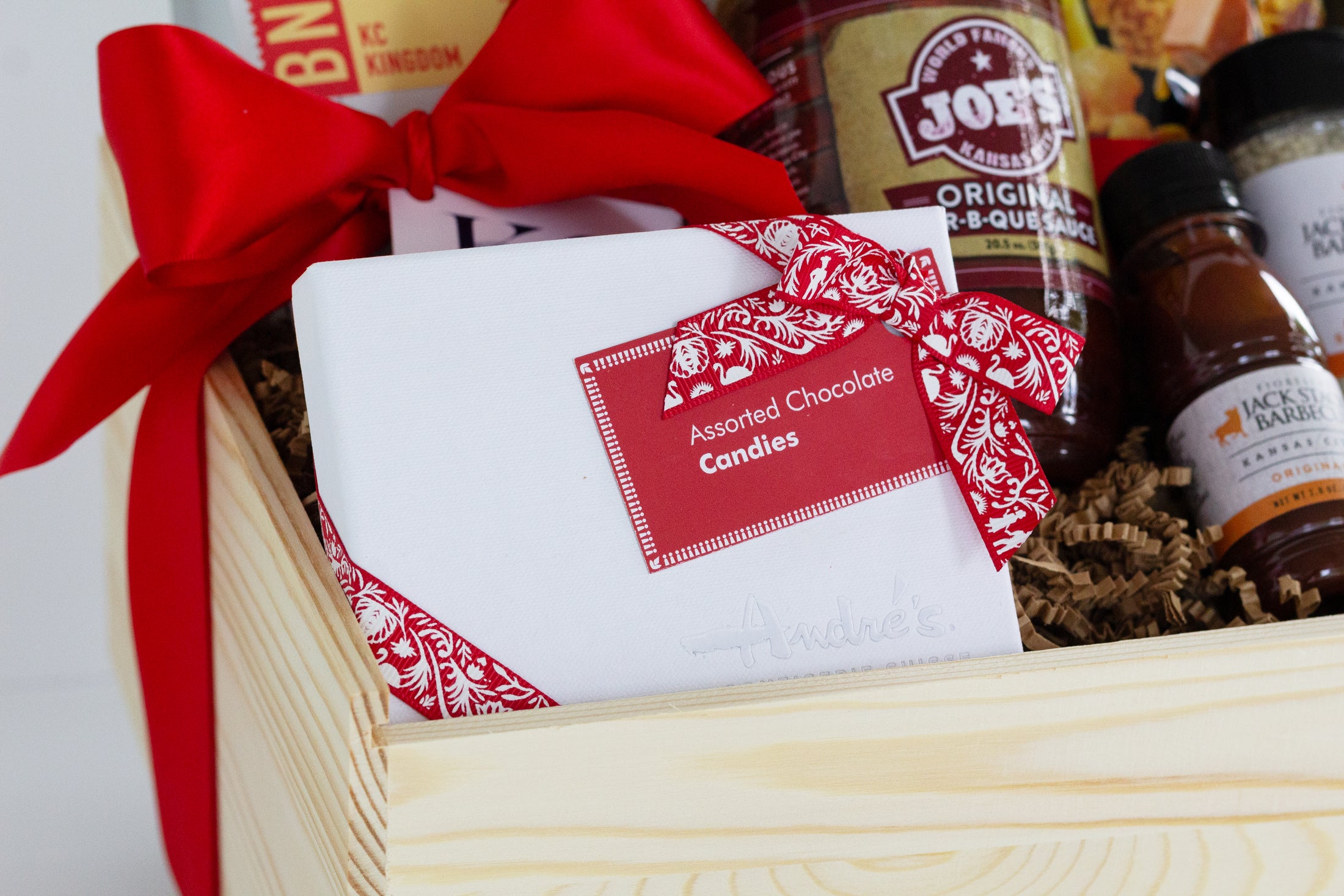 A wooden gift basket filled with coffee and tea items, including Andre's Chocolates, a bag of Topsy's Popcorn KC Mix, Roasterie Coffee, Joe's BBQ Sauce, and Jack Stack mini seasoning and bbq sauce set, arranged in a light wood crate with a red ribbon on a white background.