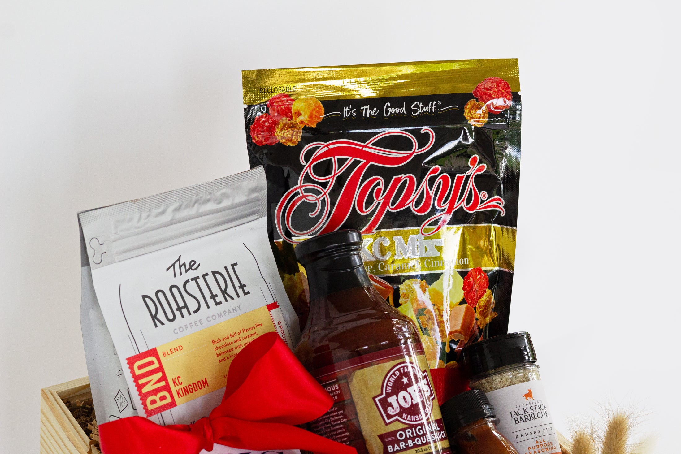 A wooden gift basket filled with coffee and tea items, including Andre's Chocolates, a bag of Topsy's Popcorn KC Mix, Roasterie Coffee, Joe's BBQ Sauce, and Jack Stack mini seasoning and bbq sauce set, arranged in a light wood crate with a red ribbon on a white background.