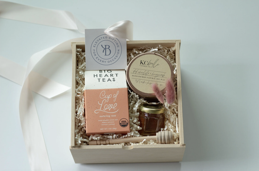 A curated gift box set containing a box of Big Heart Tea, a jar of honey, a honey stirrer, and a candle, arranged in a light wood box with shredded paper filler and a white ribbon.