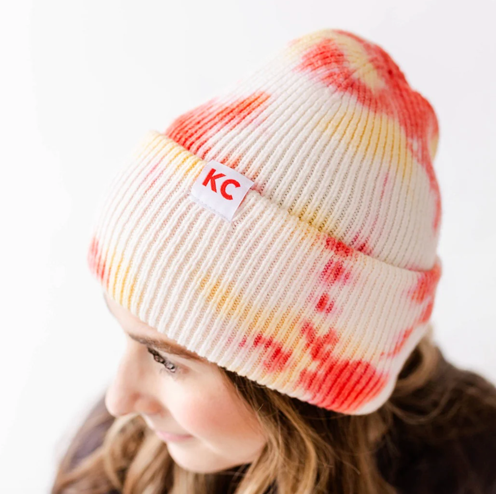 A woman with brown hair wears a red, yellow and white tie dye beanie with red "KC" on a label on the side