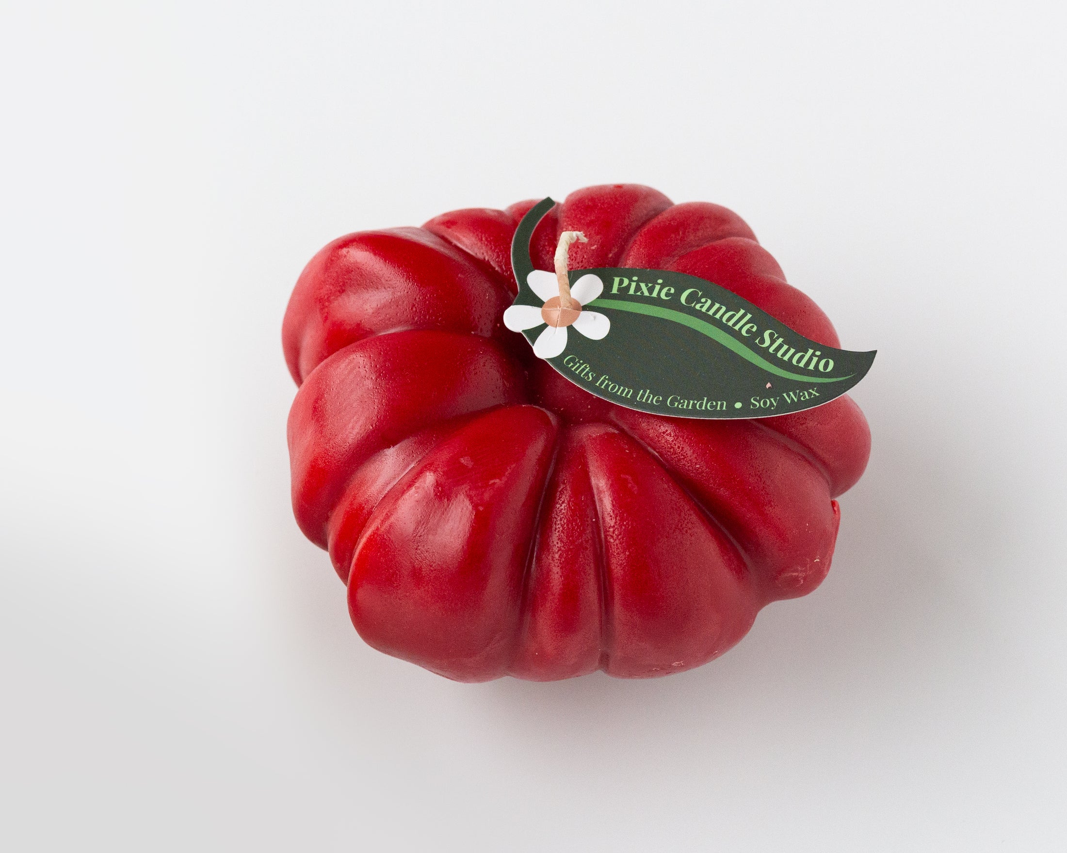A red, tomato-shaped wax candle with a dark green paper label that reads 'Pixie Candle Studio' and features a small white flower accent, set against a plain white background.
