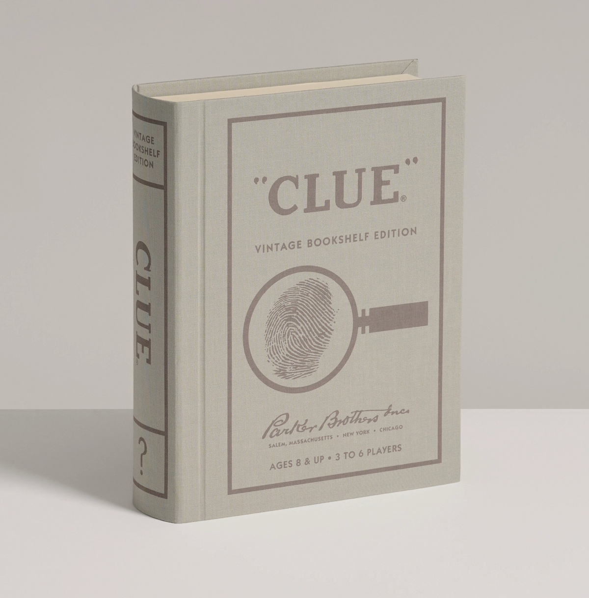 A vintage-style bookshelf edition of the board game Clue, shown standing upright on a neutral light gray background. The book features a gray cover with brown printed text and a magnifying glass fingerprint graphic.