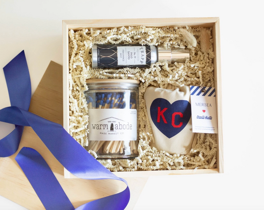 A small wood crate gift box with ivory colored krinkle paper is filled with a MERSEA Co Charlie Hustle KC Heart Candle, a glass jar of blue-tipped matches that reads WARM ABODE, and a palo santo room spray from TRAPP Candle Company. There is a blue gift ribbon and the wood top to the wood crate also in frame. 