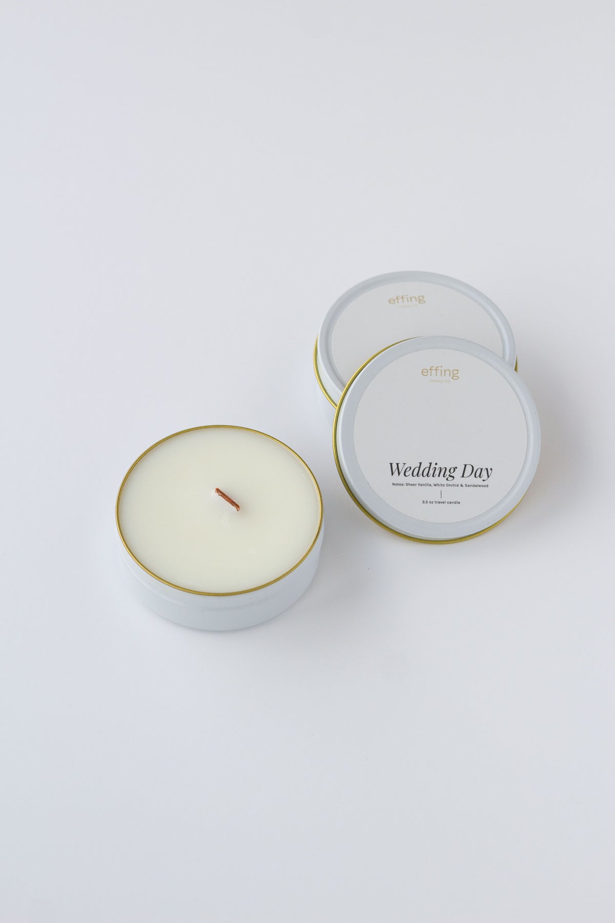 Two round candles in white metal tins with gold rims on a clean white background. One tin is open, showing pale cream wax and a single wood wick, while the other one is closed with white labels reading 'effing candle co' and 'Wedding Day'.