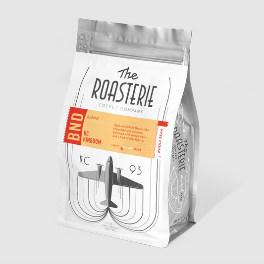 A stand-up coffee bag from The Roasterie Coffee Company, featuring a white package with black typography, a red-orange BND label, and a stylized airplane graphic on a light gray background.