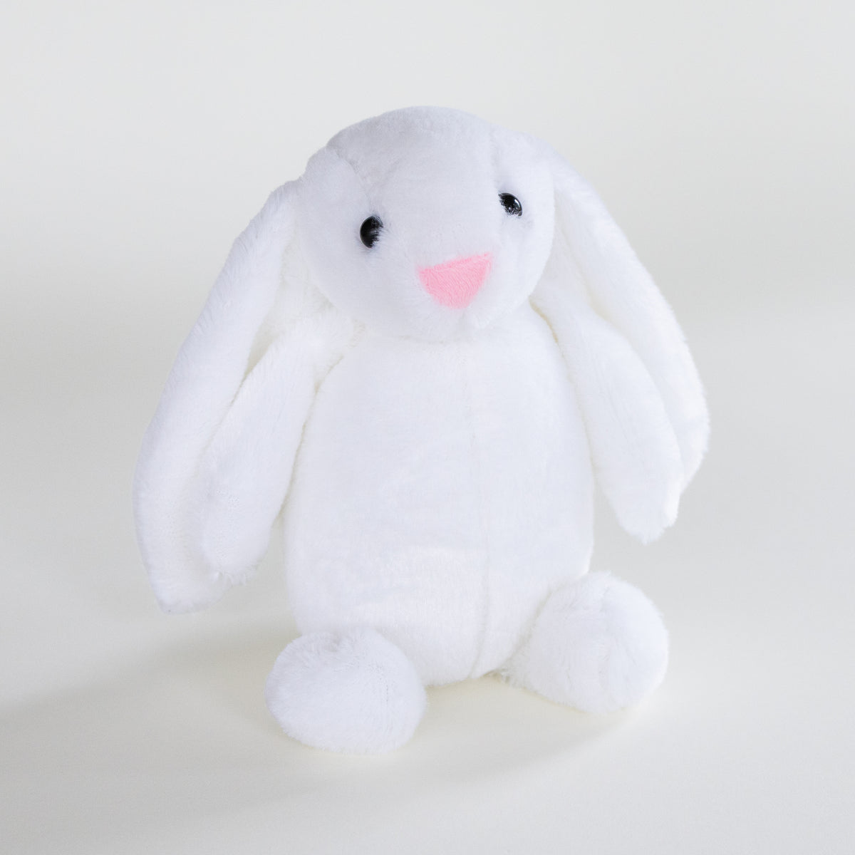 A white plush bunny toy with long floppy ears, small black eyes, and a pink stitched nose, sitting against a plain light background.