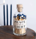 Load image into Gallery viewer, A clear glass bottle with a cork stopper filled with wooden matchsticks and blue match heads, labeled 'PEACE and LIGHT,' sitting on a dark wooden surface with blurred navy candles in the background.
