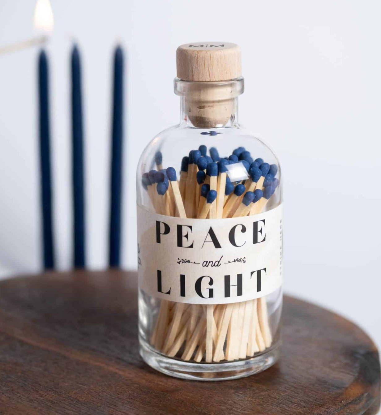 A clear glass bottle with a cork stopper filled with wooden matchsticks and blue match heads, labeled 'PEACE and LIGHT,' sitting on a dark wooden surface with blurred navy candles in the background.