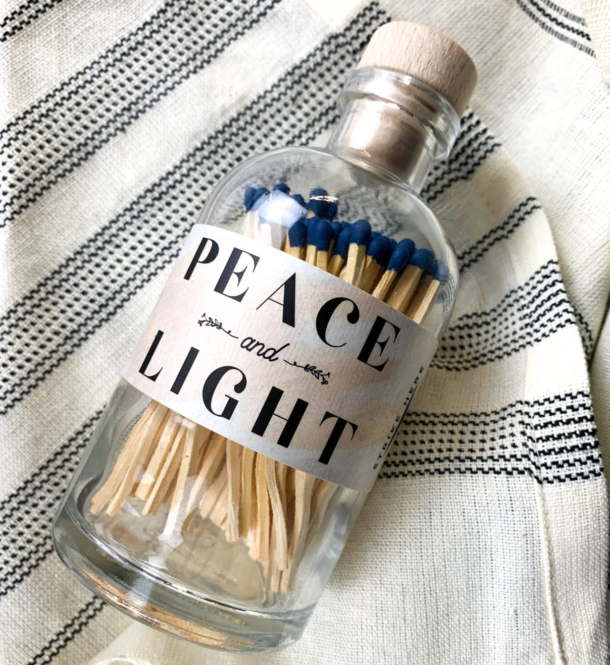 A clear glass bottle filled with wooden matchsticks, sealed with a light wood cork, and labeled 'PEACE and LIGHT' in black text. The bottle is placed on a cream fabric with black woven stripes.
