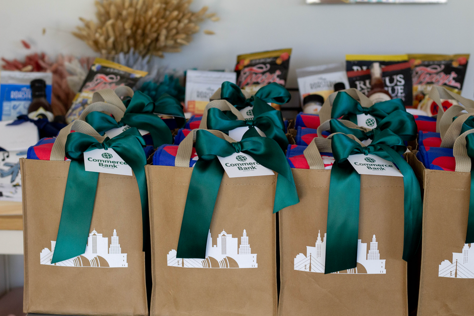 A row of brown paper gift bags tied with large green ribbons and bows, each labeled Commerce Bank, arranged on a table with assorted packaged items in the background.