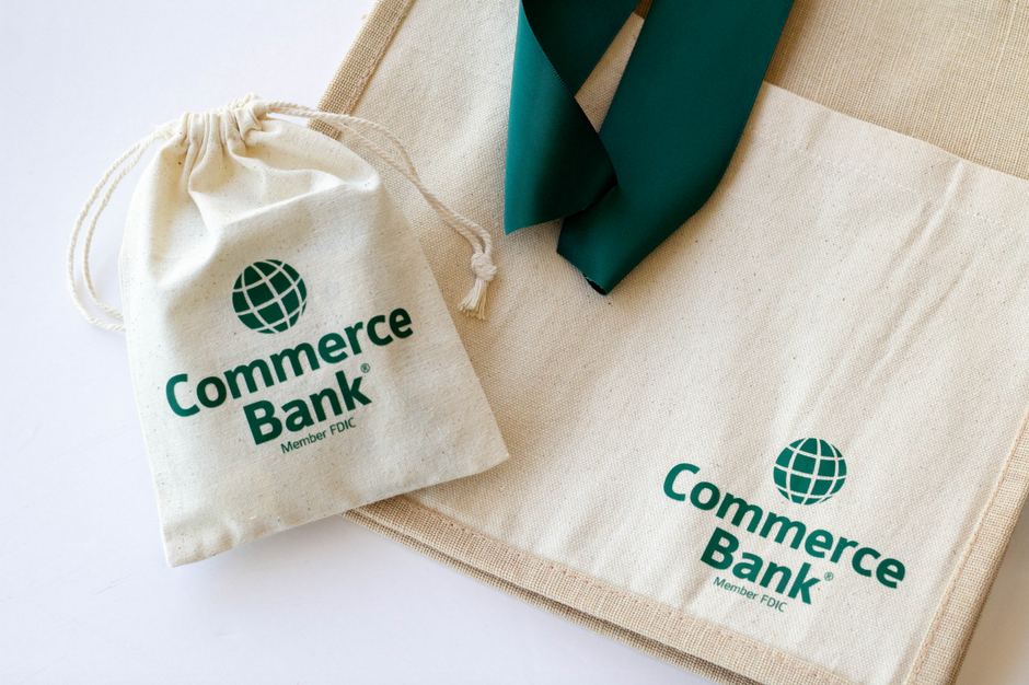 A beige canvas drawstring pouch and matching tote bag printed with the green Commerce Bank logo, with a dark green ribbon draped across the tote, on a white background.