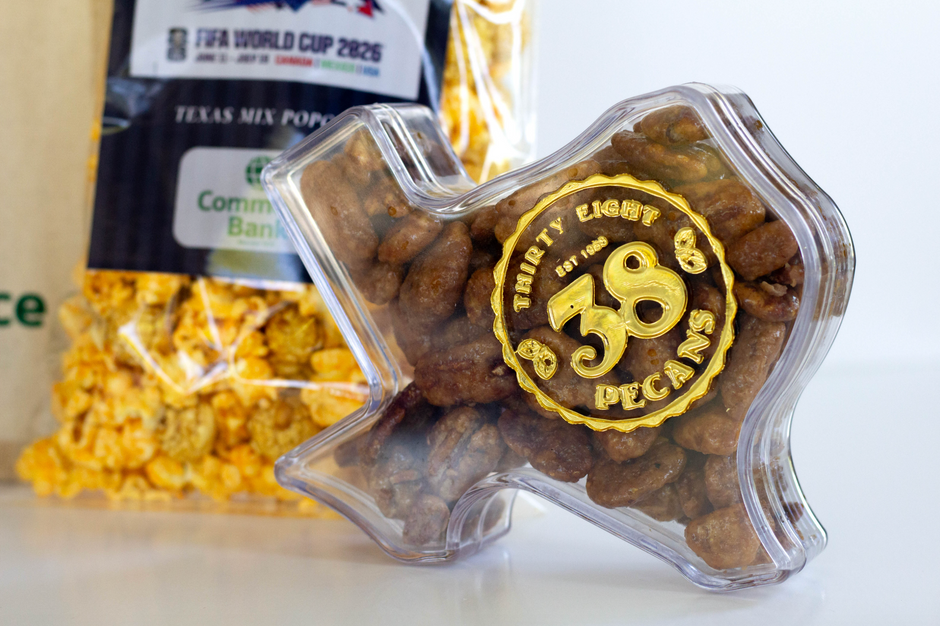 A clear plastic container shaped like the state of Texas holds roasted pecans with a gold circular label reading 'Thirty Eight Pecans Est. 2026.