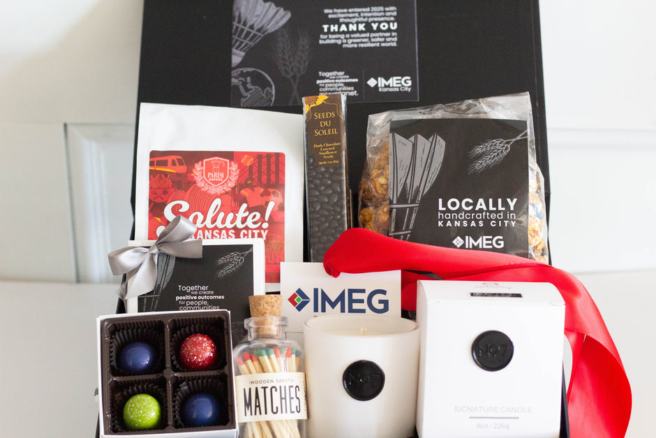 A curated gift basket filled with multiple items including IMEG coffee, chocolate covered sunflower seeds, a popcorn mix, a white candle, a box of multi-colored chocolates in IMEG branding, matches, and a red ribbon, set against a plain light background.