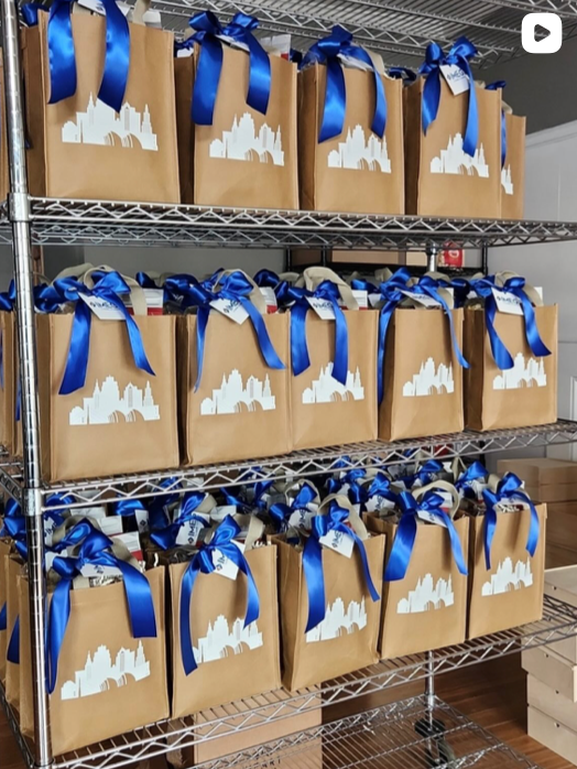 Multiple brown paper gift bags with white city skyline cutouts and bright blue ribbons tied at the top, arranged on metal wire shelves.