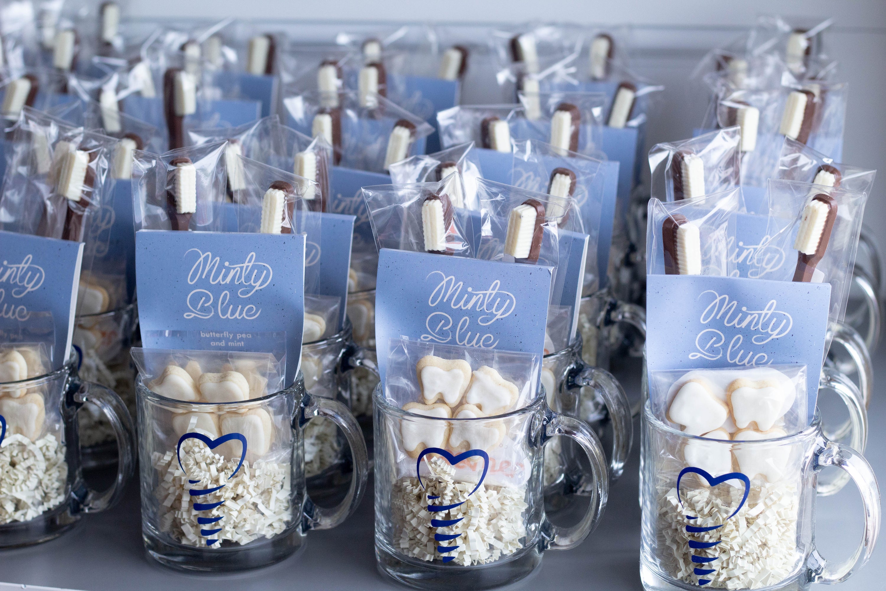 A row of packaged treats in branded glass coffee mugs with blue tea bags reading 'Minty Blue,' miniature white frosted cookies shaped like teeth, and chocolate and white chocolate toothbrushes. 