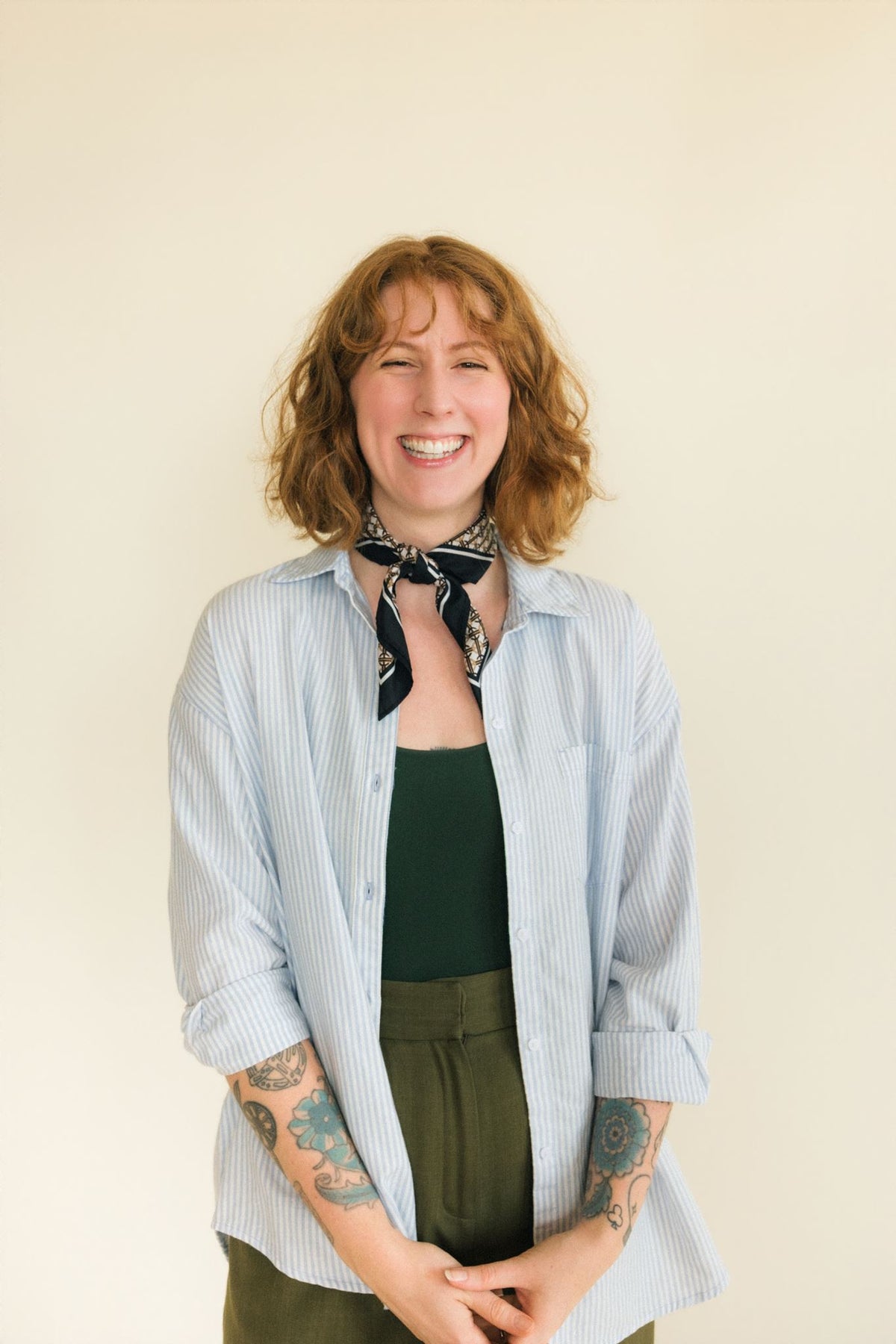 A smiling woman with shoulder-length wavy light red hair stands against a plain cream background. She is wearing a light blue and white vertically striped button-up shirt with rolled sleeves, a dark green tank top, olive green pants, and a black patterned neck scarf.