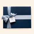 Load image into Gallery viewer, A navy blue high quality cardboard gift box wrapped with a light blue satin ribbon and bow, featuring a white tag with a circular logo.
