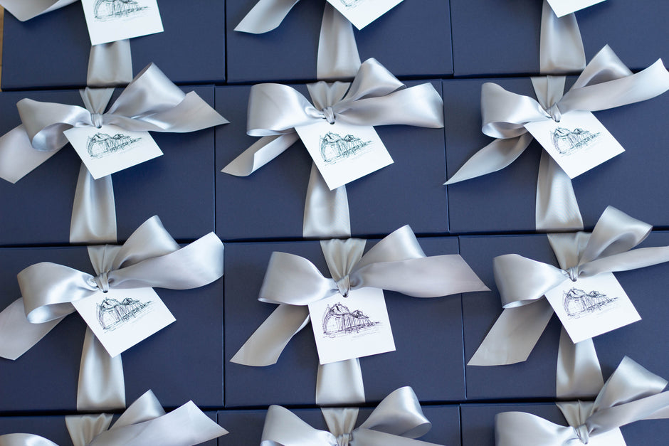 A close-up of multiple dark navy gift boxes arranged in a grid, each tied with a white satin ribbon and a small white tag printed with a delicate illustration.