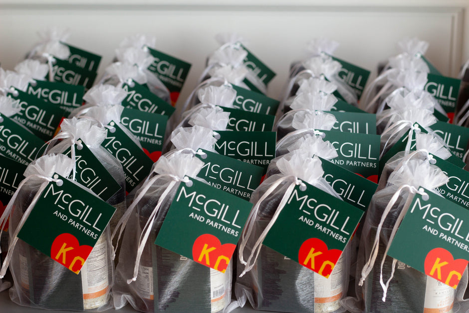 Multiple small gift bags tied with white ribbon, each featuring a dark green McGill and Partners tag, arranged in rows.