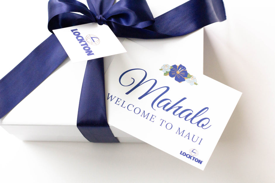 A white gift box tied with a navy blue satin ribbon and bow, featuring a white tag that reads 'Mahalo Welcome to Maui' and a smaller tag that reads 'Locton'.