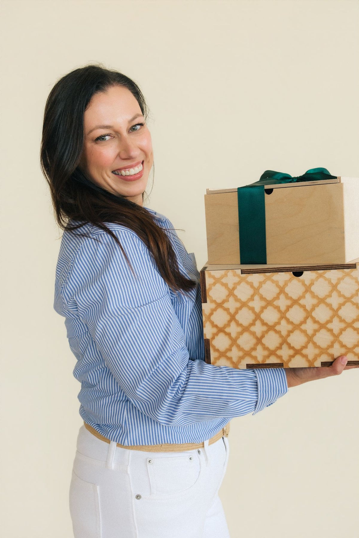 A smiling woman with long dark hair, wearing a blue and white striped shirt and white pants, holds two wooden storage boxes against a plain beige background. One box is plain natural wood with a dark green ribbon, and the other has a tan geometric trellis pattern.