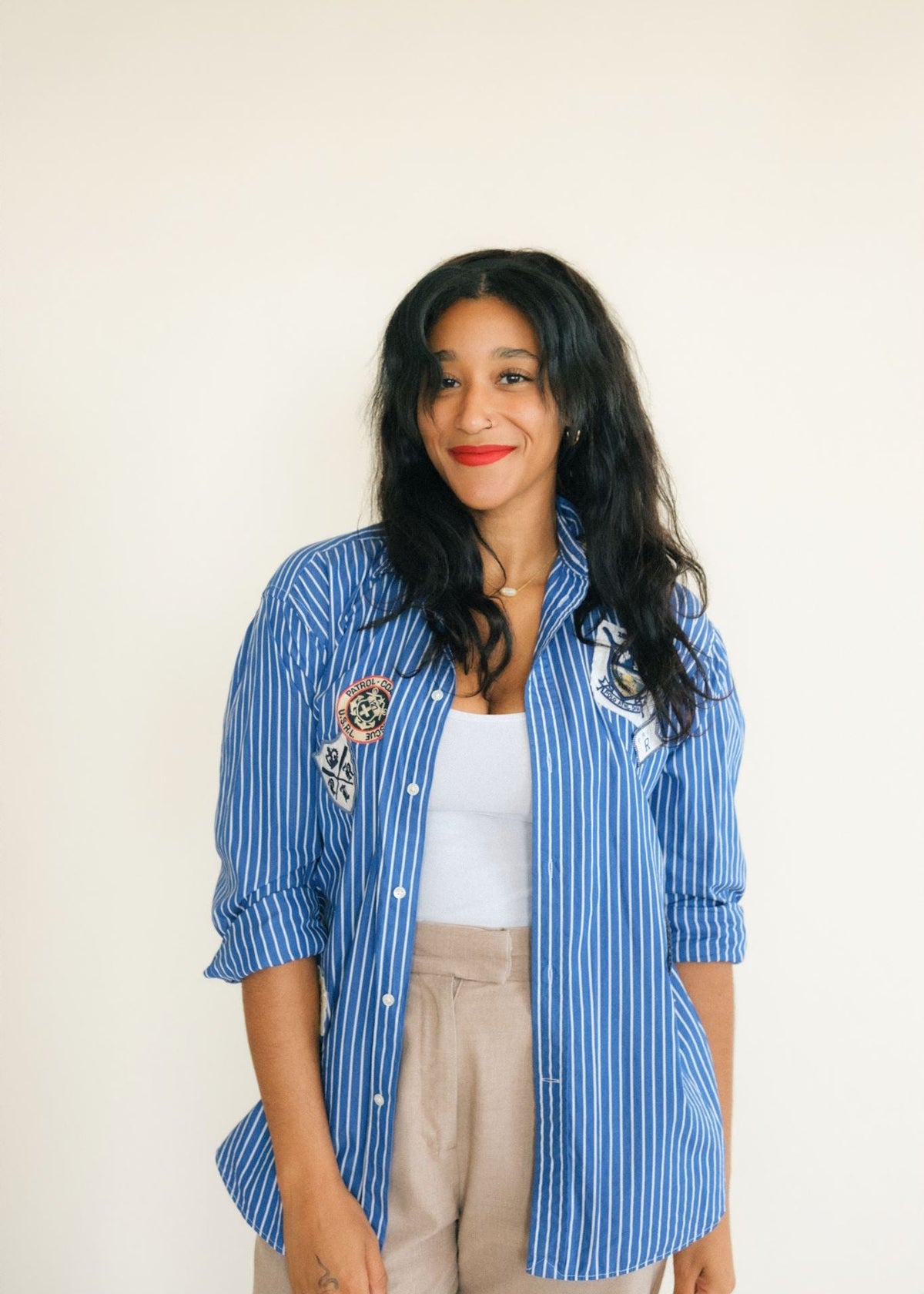 A woman with long dark hair stands against a plain off-white background wearing a blue and white vertically striped button-up shirt with embroidered patches on the chest, a white tank top, and beige pants.