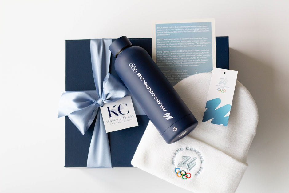 A navy blue insulated water bottle labeled 'Milano Cortina 2026' for the Winter Olympics 2026, atop a navy blue gift box with light blue ribbon and a white beanie embroidered with the 2026 Winter Olympics logo.