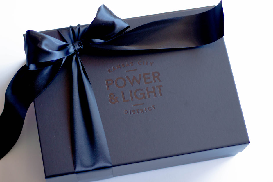 A dark navy blue gift box tied with a matching satin ribbon, featuring the embossed text 'POWER & LIGHT DISTRICT' and 'KANSAS CITY' on a clean white background.