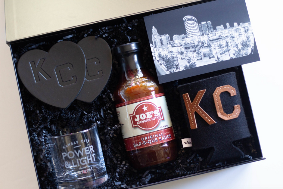 A curated gift box set featuring a bottle of Joe's Kansas City Bar-B-Que Sauce, a clear glass with 'POWER & LIGHT' printed on it, two dark gray heart-shaped coasters with 'KC' lettering, a black fabric can cooler with 'KC' leather patches, and a black card with a black-and-white city skyline.