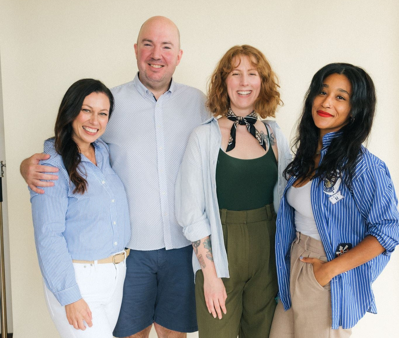 Four people standing together in a studio setting, wearing casual outfits. The group includes a man in a light blue button-down shirt and navy shorts, a woman in a dark green top with abutton down and wide-leg pants, and two women in blue striped button-down shirts with beige pants and orange shoes.