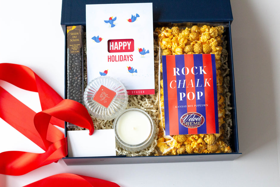 A curated gift box set on a white background, featuring a white 'Happy Holidays' card with blue birds, a pack of 'Rock Chalk Pop' popcorn, a MERSEA Co. candle, and chocolate-covered sunflower seeds, all arranged in a navy blue box with shredded paper filler and a red ribbon.