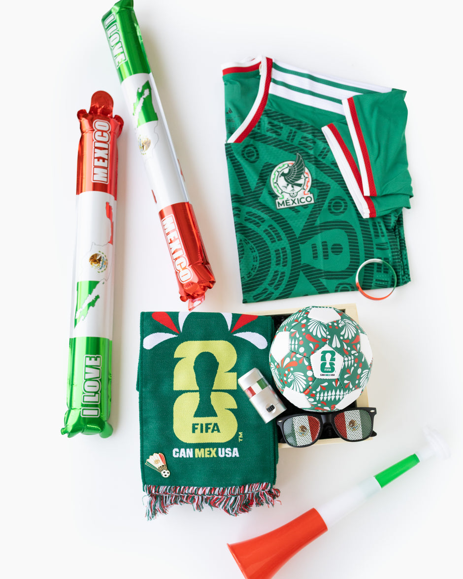 A flat lay of Mexico soccer merchandise on a white background, including a green jersey with Mexican flag trim, two inflatable noisemakers, a scarf, a soccer ball, sunglasses, a small bottle, and a horn.
