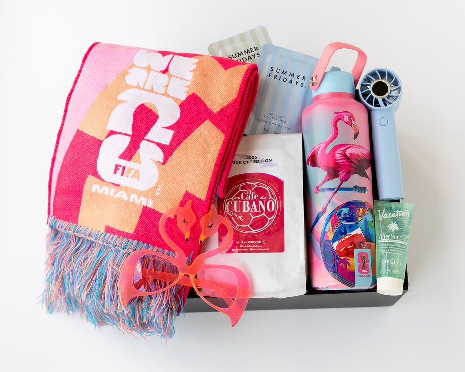 A curated gift box containing a pink and orange Miami-themed scarf, a pink flamingo water bottle, a light blue handheld fan, a tube of Vacation After Sun gel, a bag of Cafe Cubano Coffee, and other summer-themed items, all arranged in a black gift box on a white background.