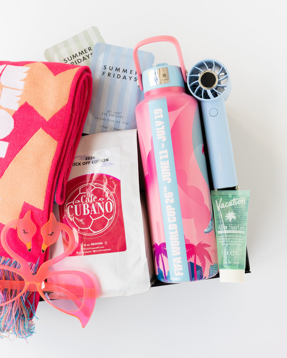 A curated gift box containing a pink and orange Miami-themed scarf, a pink flamingo water bottle, a light blue handheld fan, a tube of Vacation After Sun gel, a bag of Cafe Cubano Coffee, and other summer-themed items, all arranged in a black gift box on a white background.