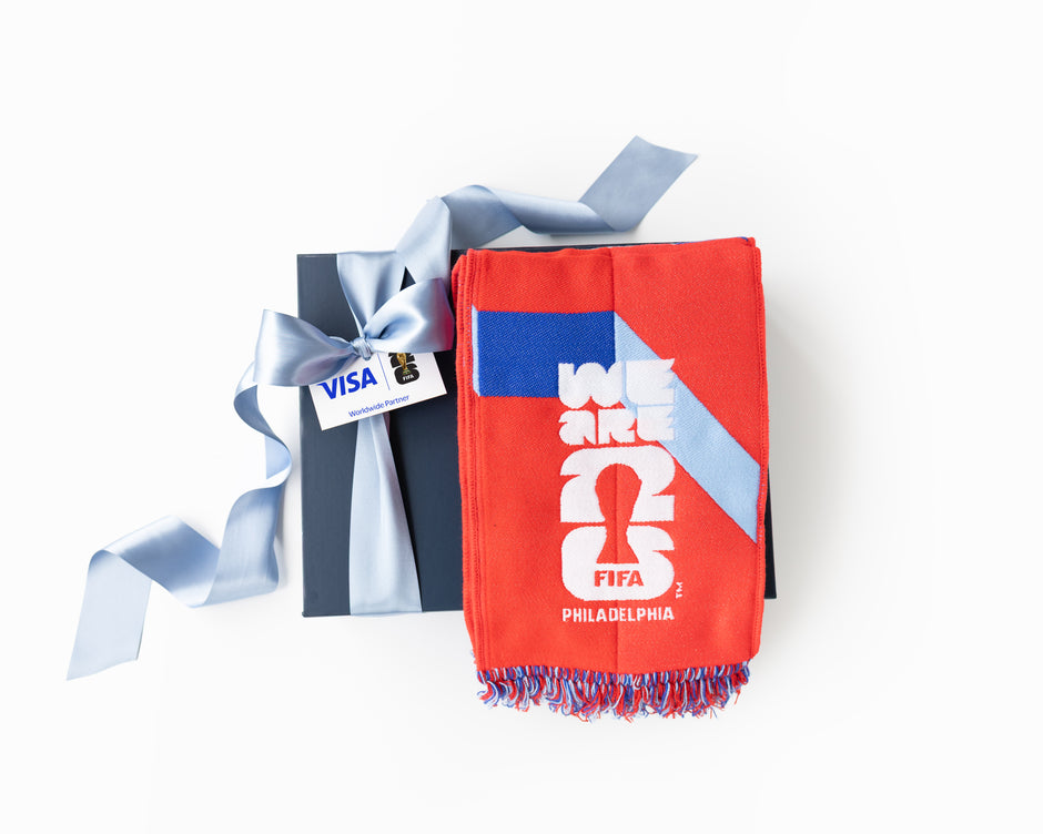 A red and blue Philadelphia soccer scarf with white graphics and fringe, displayed next to a dark gray gift box tied with a light blue ribbon. The scarf features the text 'WE ARE 2026 FIFA PHILADELPHIA'.